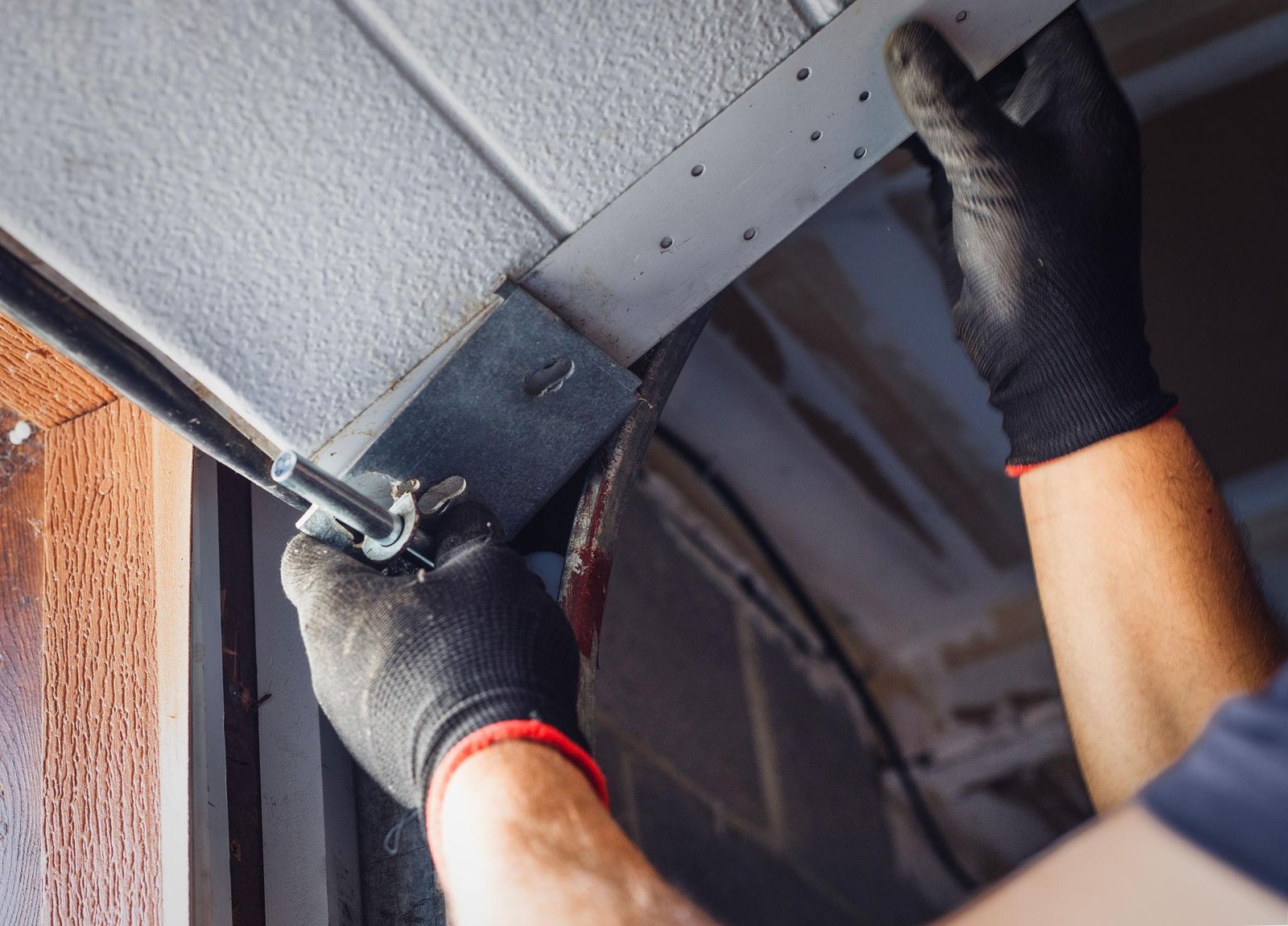 Technician tightening a metal bracket on a garage door track with gloved hands.