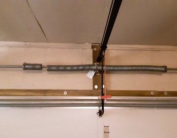 Mounted torsion spring and shaft above a garage door with central bracket and red handle.